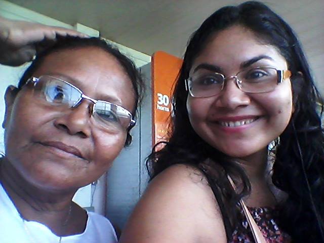 Na foto duas mulheres negras, mNa foto, duas mulheres negras, Mãe Anna e sua filha Kathellem, estão lado a lado. Ambas usam óculos e sorriem levemente. Mãe Anna está com a mão direita na cabeça.ãe Anna e sua filha Kathellem, as duas usam oculos e sorriem levemente e estão lado a lado, mãe Anna está com a mãe direita na cabeça