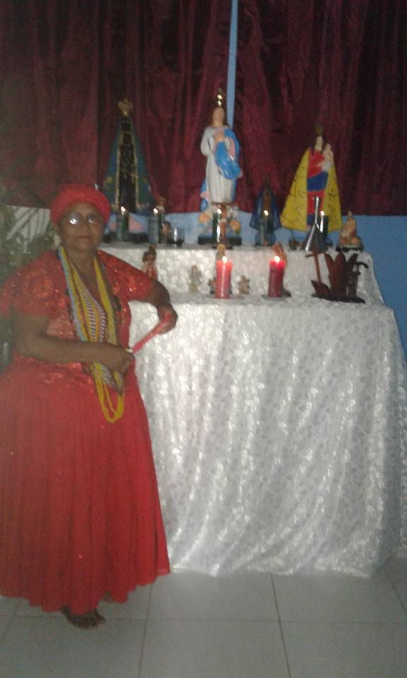 Mãe Anna está em frente a seu Altar Minutos antes de iniciar seus trabalhos, com indumentária da religião de Matriz Africana, um vestido longo vermelho e um pano de cabeça também vermelho do mesmo tecido do vestido, muitas guias em seu pescoço. Mãe Anna apoia o braço esquerdo no altar que está coberto por um longo tecido branco bordado com três imagens , uma de Nossa Senhora da Conceição, outra de São Lázaro e uma outra de nossa senhora de Nazaré e a esquerda uma imagem pequena de Yansã. Há muitas velas de 7 dias acesas.