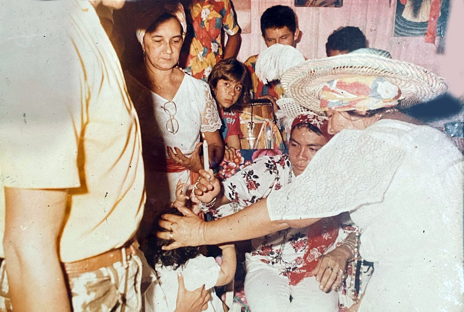 A imagem é uma foto antiga digitalizada e registra uma cerimônia religiosa, ao centro há um homem com uma blusa de botão florida e um pano de cabeça com a mesma estampa segurando uma vela acima da cabeça de Kathellen que é uma criança de no máximo 2 anos, vestidas de branco. Ao redor tem 6 pessoas, duas mulheres vestidas de branco com mangas rendadas e com panos de cabeça. Uma criança ao fundo com olhar de curiosidade também chama a atenção na imagem.