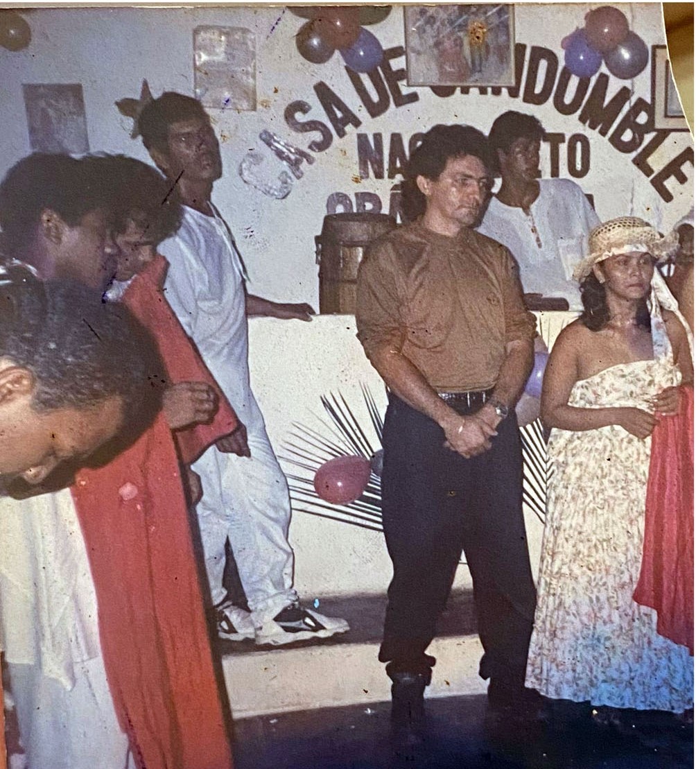A imagem é uma fotografia antiga digitalizada, na lateral 4 homens vestidos de branco, ao centro um homem aparentando meia idade vestido com uma blusa marrom e calça preta, ao seu lado Mãe Anna, mulher de meia idade com um vestido florido estilo tomara que caia e um chapéu de palha com um lenço vermelho na mão. Ao fundo uma parede branca escrito Casa de Candomblé, umas palhas, alguns balões vermelhos e um atabaque.