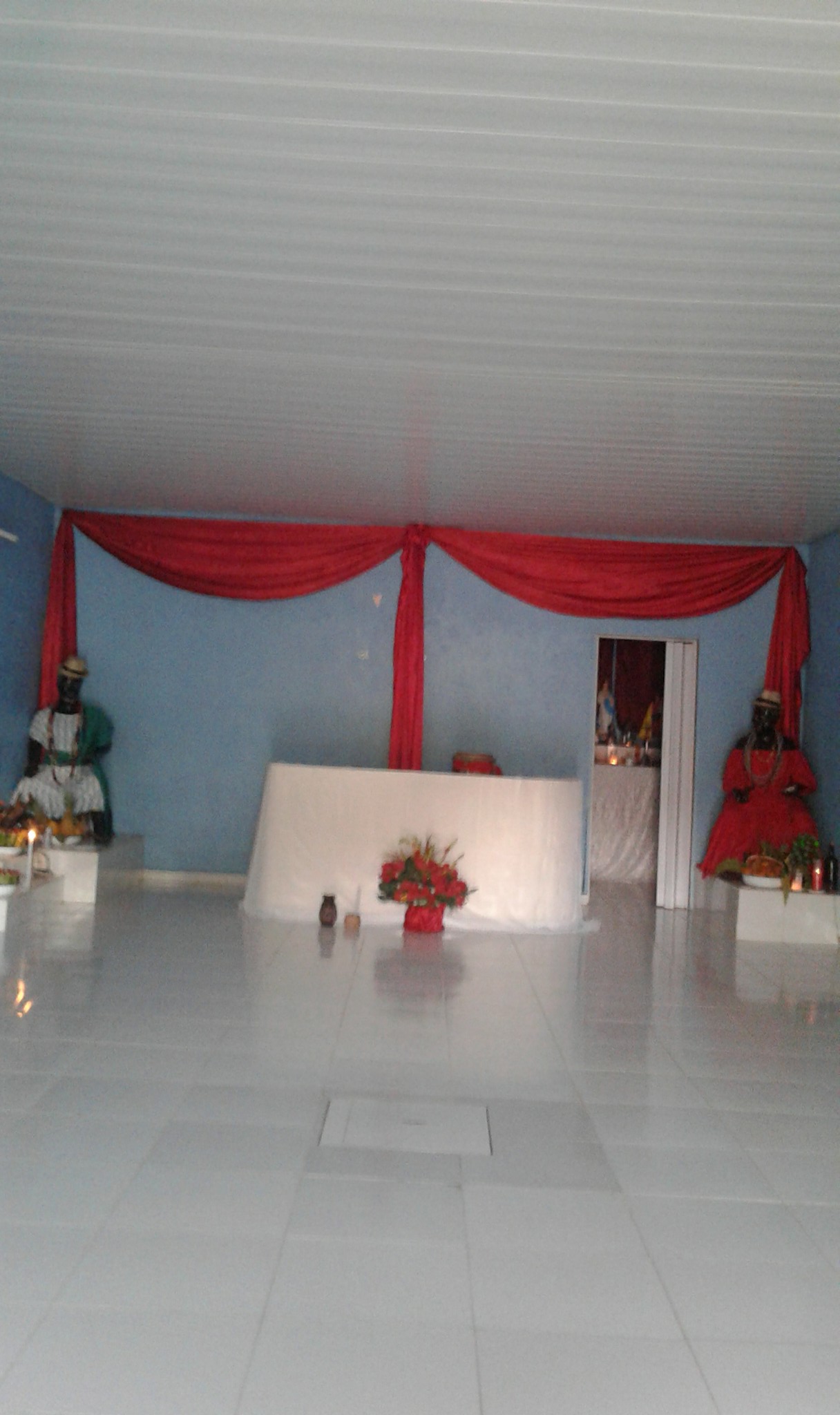 Na fotografia o registro do Terreiro de Mãe Anna, um salão aberto, na parte superior aparece o forro branco, na parte central o Altar coberto por um pano branco, a frente um buquê de rosas vermelhas, uma parede lisa azul decorada com tecidos vermelhos. Nas laterais ao fundo há duas imagens sagradas, uma masculina vestida de branco, com lenço verde e chapéu de palha e uma feminina com vermelho e um chapéu de palha. A maior parte da imagem se concentra em mostrar o lugar aberto, então boa parte da imagem é composta pelo piso branco e a sensação de um lugar amplo.