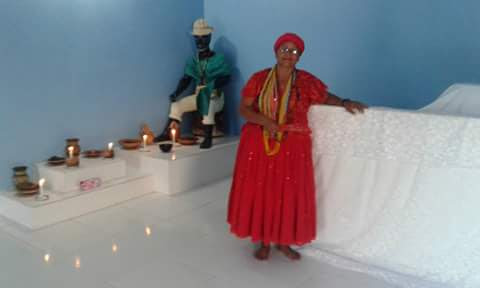 Na fotografia aparece Mãe Anna, com aparência de 40 e poucos anos, está com indumentárias de religião de Matriz Africana, uma saia e uma blusa vermelha vibrante no mesmo tom, com um pano de cabeça no mesmo tom e tecido de sua roupa. No pescoço muitas Guias, está de óculos e batom vermelho vibrante. Ela está se apoiando em 3 atabaques que estão cobertos por um longo tecido branco bordado, ao fundo, na parte esquerda da fotografia aparece uma imagem de um Preto Velho num altar, com uma blusa verde e uma calça branca, a frente da imagem algumas velas.