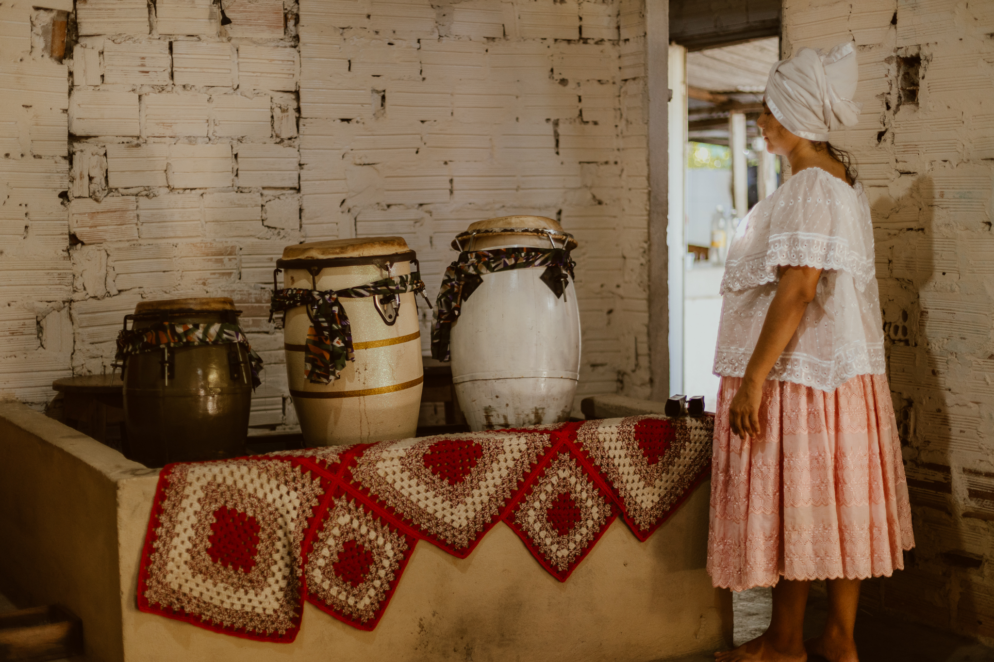 Na fotografia, em estilo paisagem, na parte direita aparece Kathellem, uma mulher negra, posicionada de frente a 3 atabaques e de lado na imagem, vestida com roupas de religião de Matriz africana. Na imagem seu corpo aparece por inteiro, ela veste uma blusa branca rendada e uma saia rosa em tom claro que vai até um pouco abaixo do joelho. Na parte esquerda os 3 atabaques, instrumentos ligados às religiões de Matriz Africana, estão em cima de uma coberta feita de crochê nas cores vermelha e branca. Ao fundo, uma parede de tijolo e uma porta que está aberta.