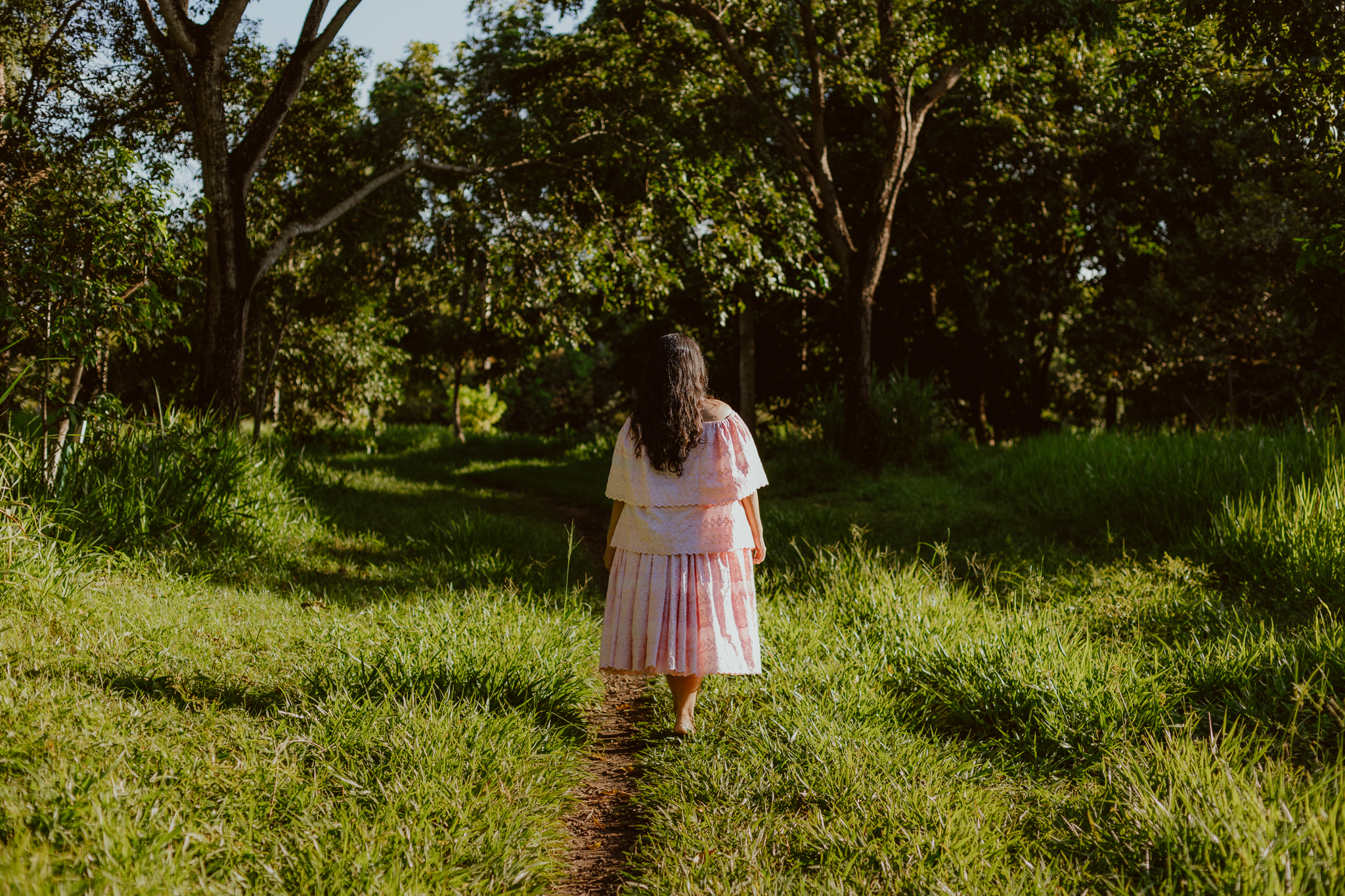 A fotografia é em close enquadrando um pouco abaixo do ombro até um pouco abaixo da cintura de Kathellen, aparece na parte superior da foto os cabelos dela somente. Kathellen veste uma blusa branca com bordados e uma saia estilo godê rosa no tom claro, na imagem aparece apenas o seu braço direito. Em seu pescoço algumas guias, uma preta e vermelha, uma verde, uma amarela, uma laranjada e uma branca.