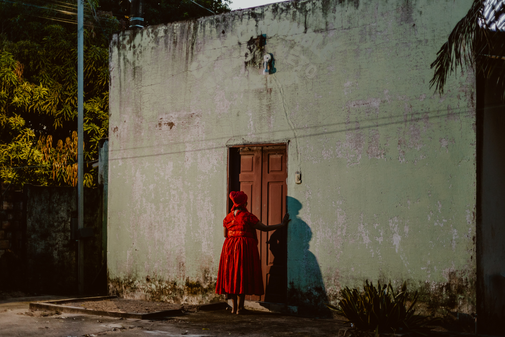 A fotografia possui um ângulo bem aberto, pegando a entrada toda do terreiro e ao centro, na frente de uma porta Kathellen , mulher negra, de estatura mediana, com indumentária de religião de matriz africana. A roupa é uma blusa e uma saia vermelha que vai até abaixo do joelho, a mesma vestimenta de Mãe Anna usa na imagem 12, e um pano de cabeça no mesmo tom vermelho cintilante. A frente do terreiro é verde desbotada, então as cores da roupa, da frente do terreiro e da porta que é marrom fazem a composição estética da imagem.