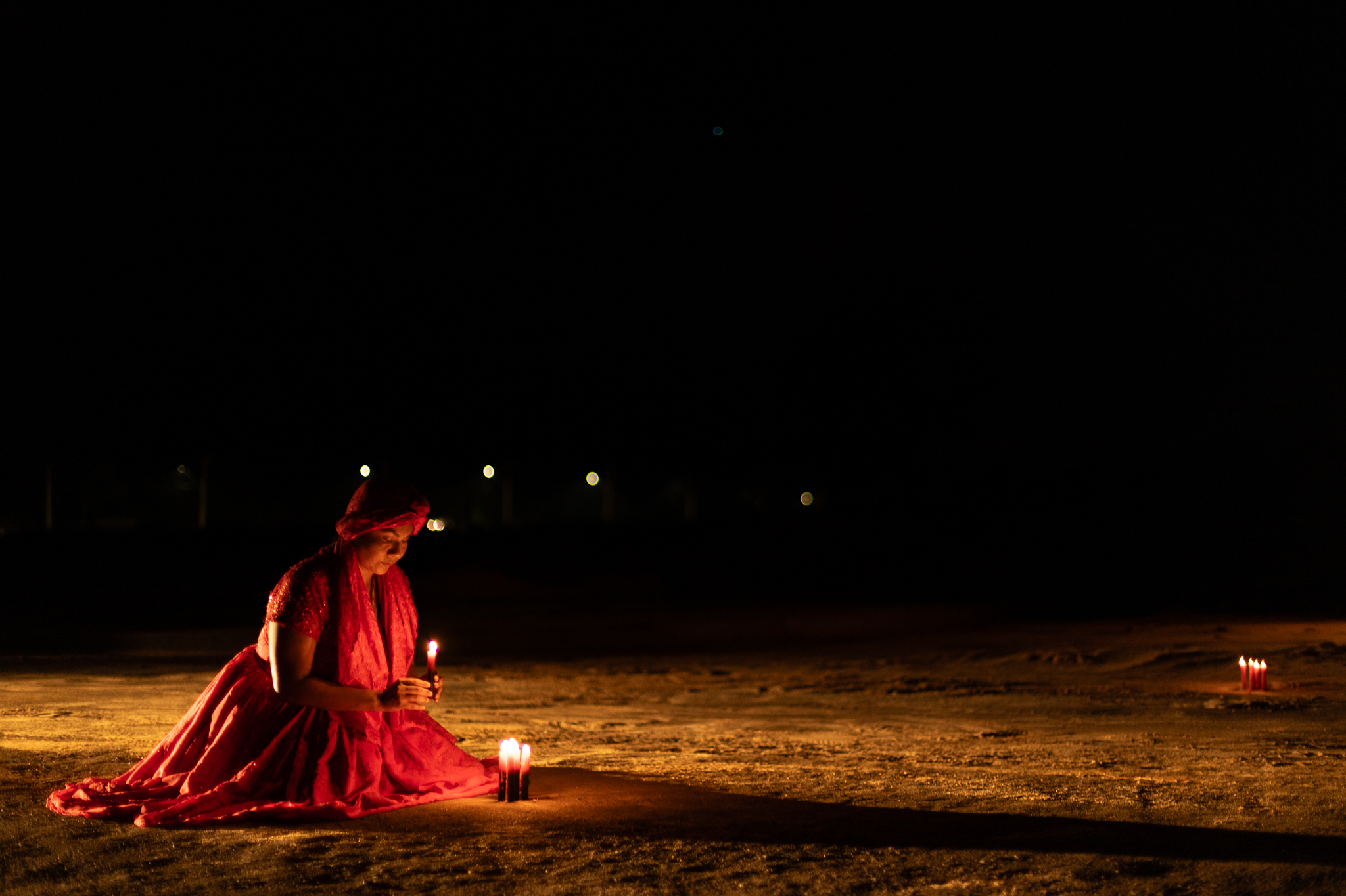 A fotografia é em estilo paisagem, uma foto noturna em que toda a parte superior é escura e na parte esquerda da imagem está Kathellen, mulher negra, que aparenta uns 28 anos, vestida com indumentária de religião de matriz Africana, a mesma saia e blusa vermelha vibrante que Mãe Anna usa na fotografia 12, com um pano de cabeça vermelhos no mesmo tom, ela está abaixada com uma vela vermelha acesa na mão e 3 velas vermelhas já estão fixadas na sua frente. Toda parte inferior da foto é de um chão praiano e ao fundo no canto direito têm-se várias velas vermelhas acesas. A composição estética da imagem se dá no jogo de luz da vela e no tom avermelhado da indumentária, contrastando com a escuridão da parte superior da foto.