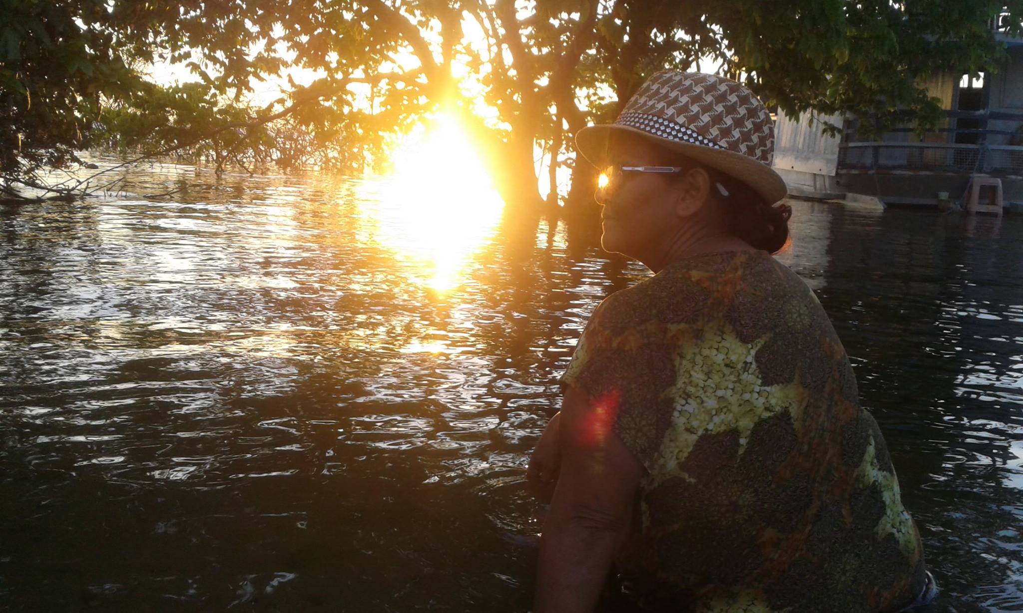 A imagem é uma fotografia em estilo paisagem, na parte direita está Mãe Anna, uma mulher que aparenta de 45 anos , com um chapéu quadriculado, óculos, e uma blusa. Ela está sentada na beira do rio, sua posição é de perfil, na parte central e esquerda da imagem o pôr do sol e as águas do rio, na parte superior galhos de árvore. Por conta da luz do sol a foto está um pouco contra luz