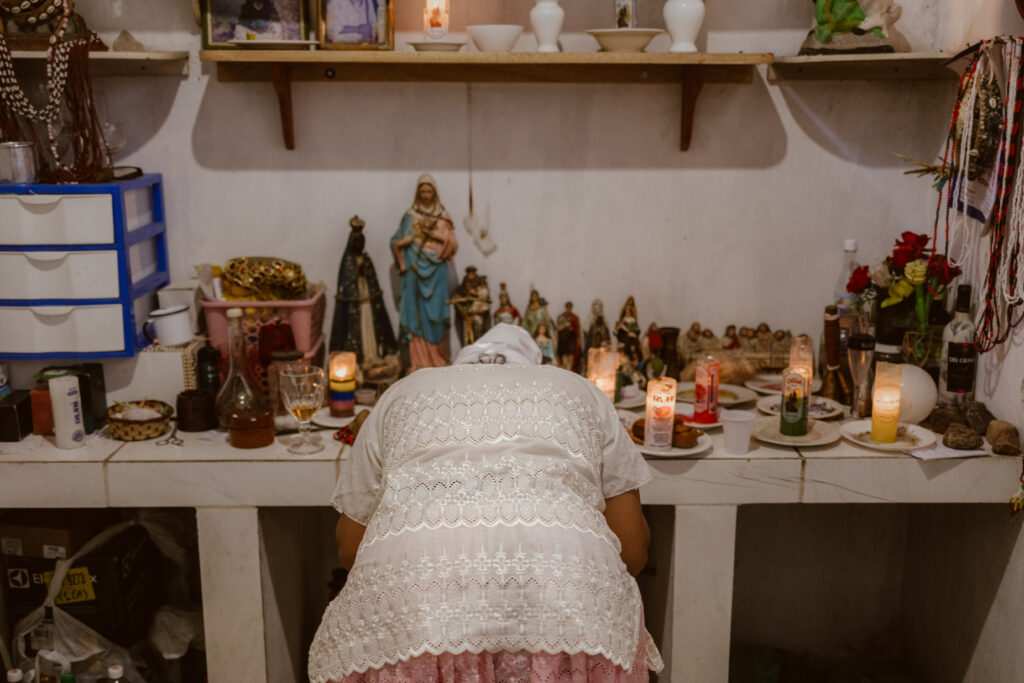 : A imagem é uma fotografia em estilo paisagem, ao centro da imagem está kathellen ajoelhada e curvada sobre um altar, não é possível vê seu rosto pois ela está posicionada de frente para o altar e de costas na imagem. O altar possui muitas velas de 7 dias acesas, imagens de santos e outras imagens pequenas, o altar ocupa a parte média da foto de ponta a ponta horizontalmente. Na parte superior da imagem é possível vê velas brancas e quartinhas brancas.
