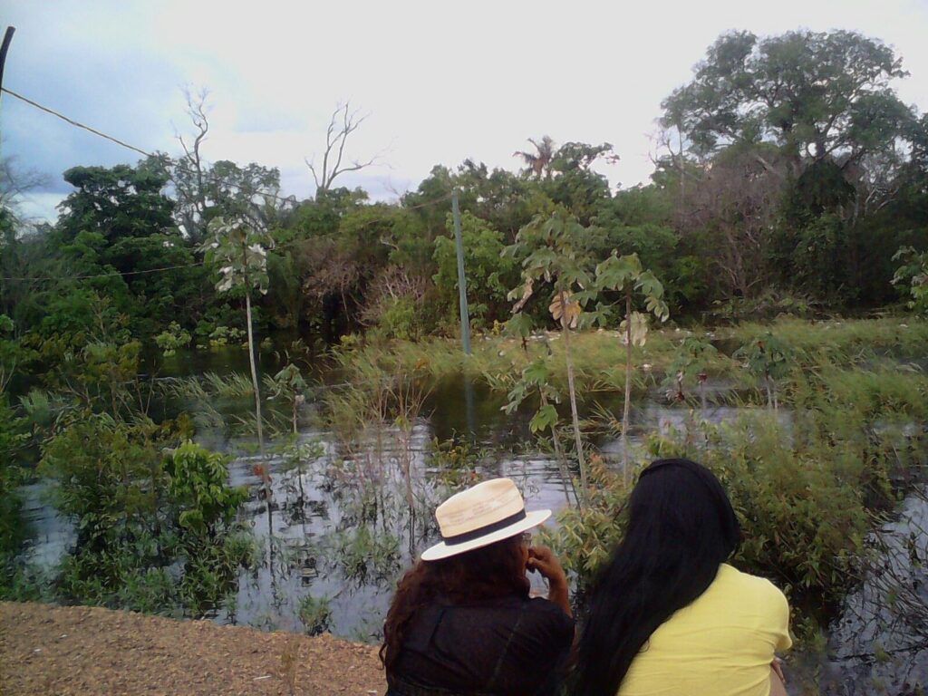 Na fotografia Mãe Anna e uma mulher estão sentadas a beira de um lago, elas aparecem de costas no primeiro plano, ao fundo o lago com água esverdeada e límpida e muitas árvores ao redor.